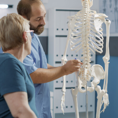 Male doctor pointing at human skeleton to show spinal cord and explain mechanical disorders in rehabilitation cabinet. Specialist explaining back bones system for physical therapy.